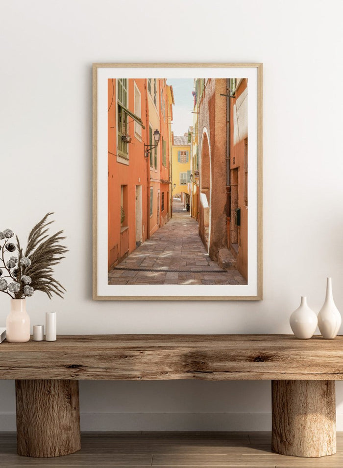 Framed photograph of a narrow street on a wall next to a window.  narrow street with colorful buildings in a European city. Vibrant European Alley by Henrike Schenk is a travel photography print featuring a Mediterranean street, cobblestone alley, and pastel European architecture.
