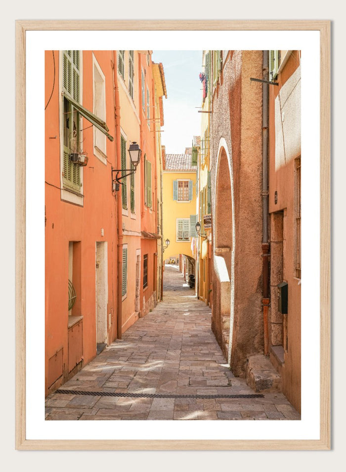Framed photograph of a narrow street on a wall next to a window.  narrow street with colorful buildings in a European city. Vibrant European Alley by Henrike Schenk is a travel photography print featuring a Mediterranean street, cobblestone alley, and pastel European architecture.