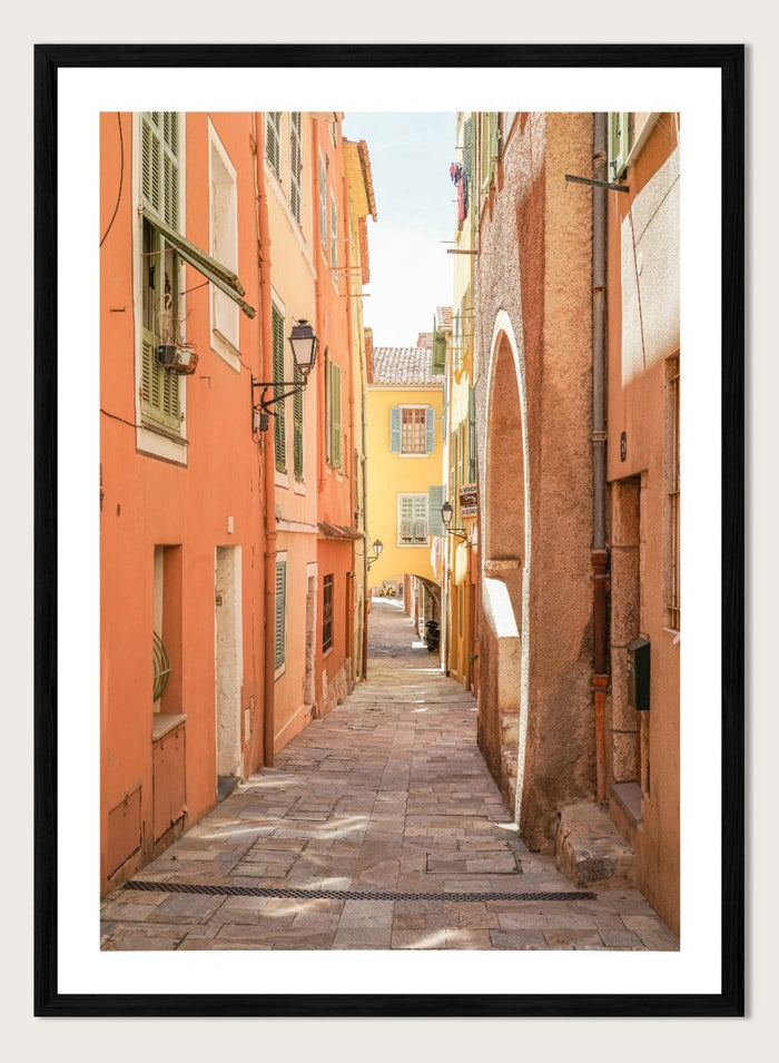 Framed photograph of a narrow street on a wall next to a window.  narrow street with colorful buildings in a European city. Vibrant European Alley by Henrike Schenk is a travel photography print featuring a Mediterranean street, cobblestone alley, and pastel European architecture.
