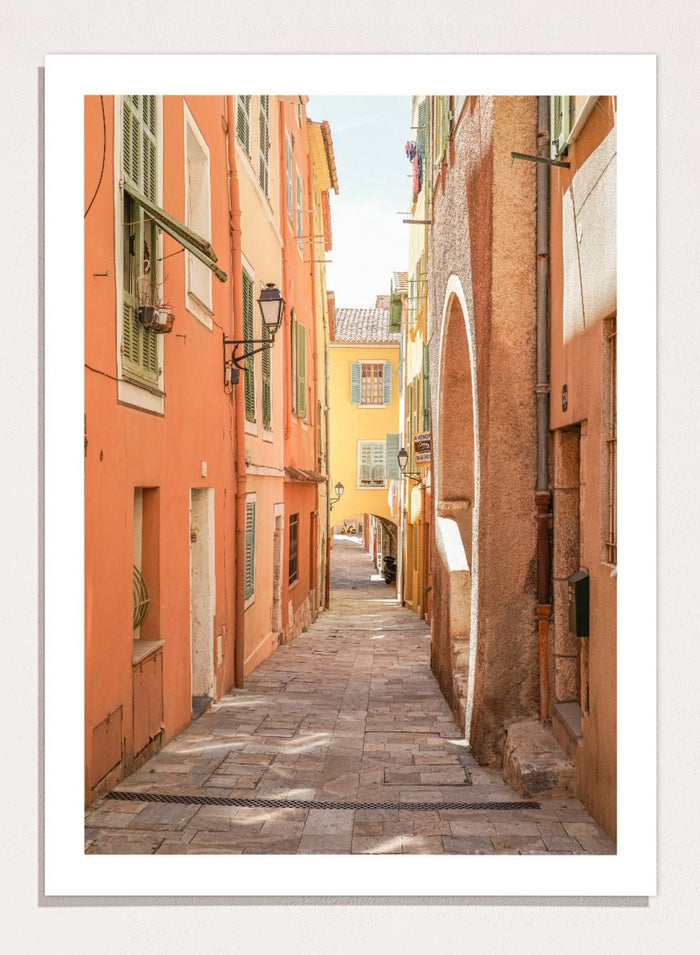 Framed photograph of a narrow street on a wall next to a window.  narrow street with colorful buildings in a European city. Vibrant European Alley by Henrike Schenk is a travel photography print featuring a Mediterranean street, cobblestone alley, and pastel European architecture.
