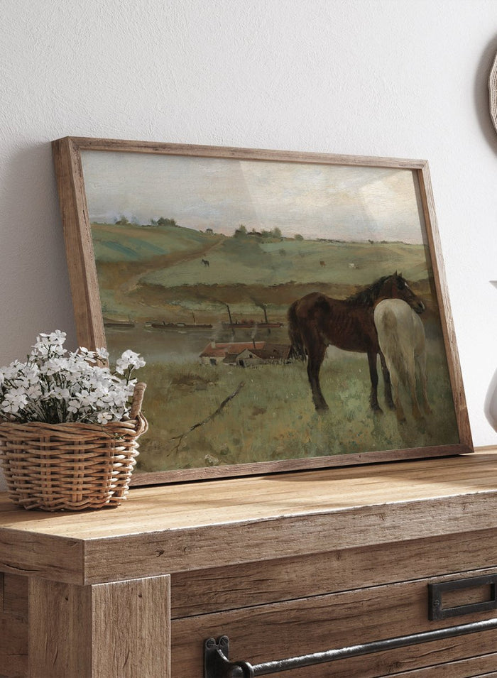 A framed landscape painting depicting two horses in a grassy field, with a muted sky and distant hills. Horses in a Meadow by Edgar Degas is a vintage horse art print featuring two horses standing together in a green field with a river, hills, and distant village.