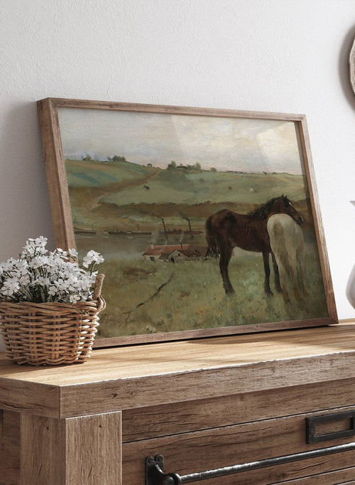 A framed landscape painting depicting two horses in a grassy field, with a muted sky and distant hills. Horses in a Meadow by Edgar Degas is a vintage horse art print featuring two horses standing together in a green field with a river, hills, and distant village.