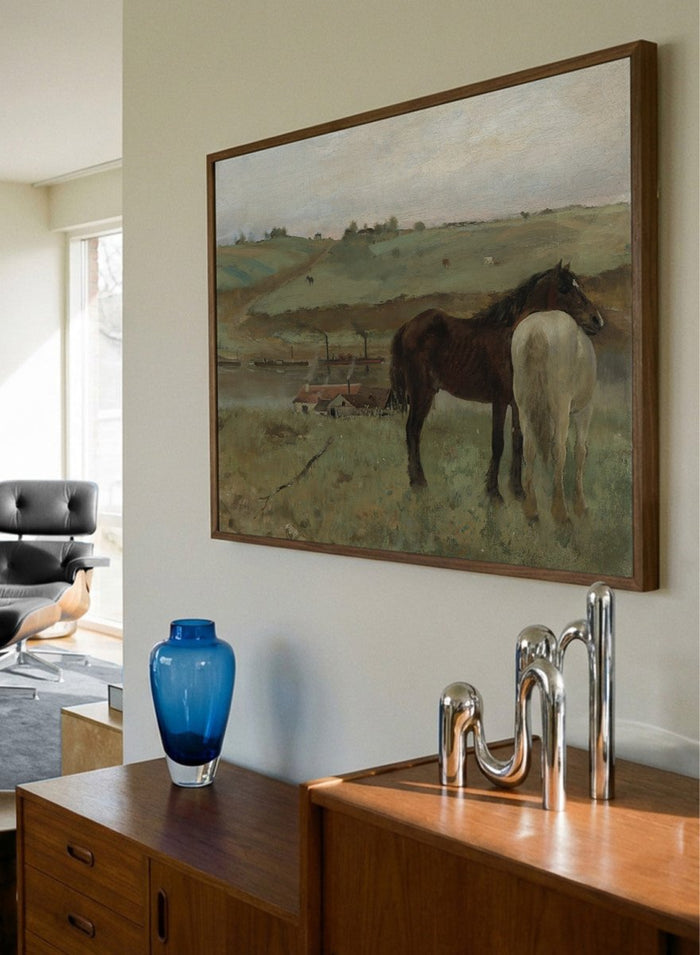 A framed landscape painting depicting two horses in a grassy field, with a muted sky and distant hills. Horses in a Meadow by Edgar Degas is a vintage horse art print featuring two horses standing together in a green field with a river, hills, and distant village.