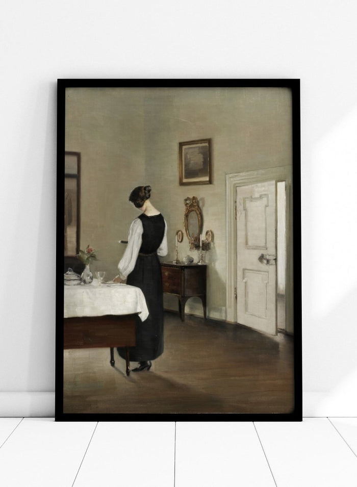 Woman in a black dress standing in a room with a table and mirror. In the Dining Room by Carl Holsøe is a vintage Danish interior art print featuring a woman in a softly lit dining room with muted tones and classic Scandinavian style.