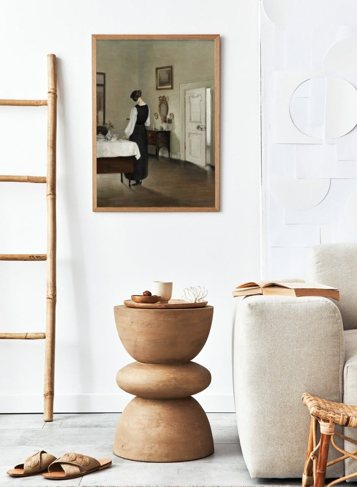 Woman in a black dress standing in a room with a table and mirror. In the Dining Room by Carl Holsøe is a vintage Danish interior art print featuring a woman in a softly lit dining room with muted tones and classic Scandinavian style.