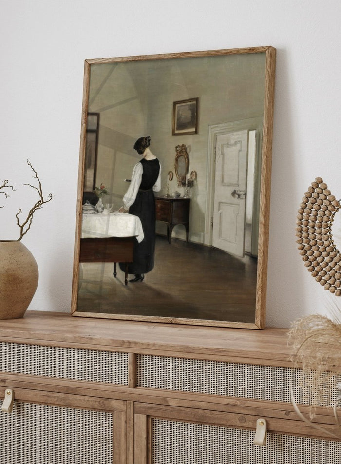 Woman in a black dress standing in a room with a table and mirror. In the Dining Room by Carl Holsøe is a vintage Danish interior art print featuring a woman in a softly lit dining room with muted tones and classic Scandinavian style.