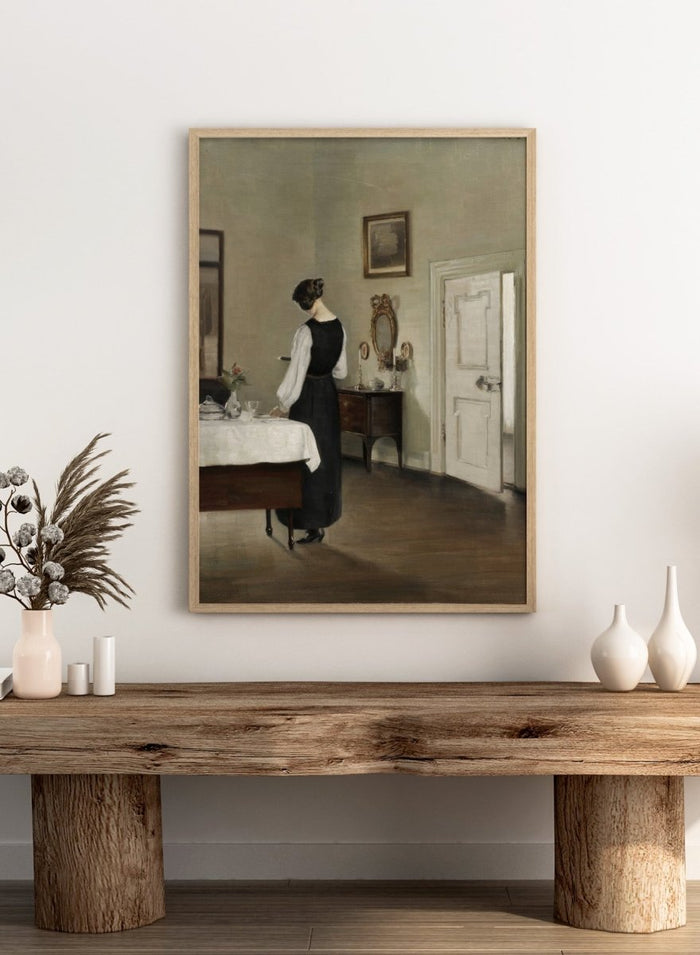 Woman in a black dress standing in a room with a table and mirror. In the Dining Room by Carl Holsøe is a vintage Danish interior art print featuring a woman in a softly lit dining room with muted tones and classic Scandinavian style.