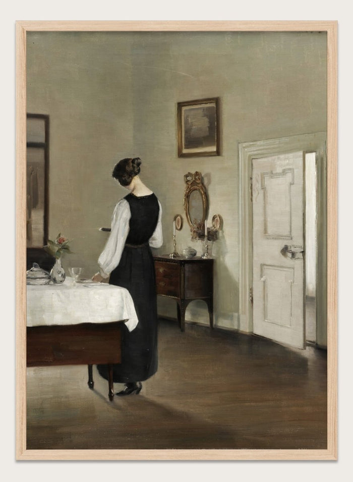 Woman in a black dress standing in a room with a table and mirror. In the Dining Room by Carl Holsøe is a vintage Danish interior art print featuring a woman in a softly lit dining room with muted tones and classic Scandinavian style.