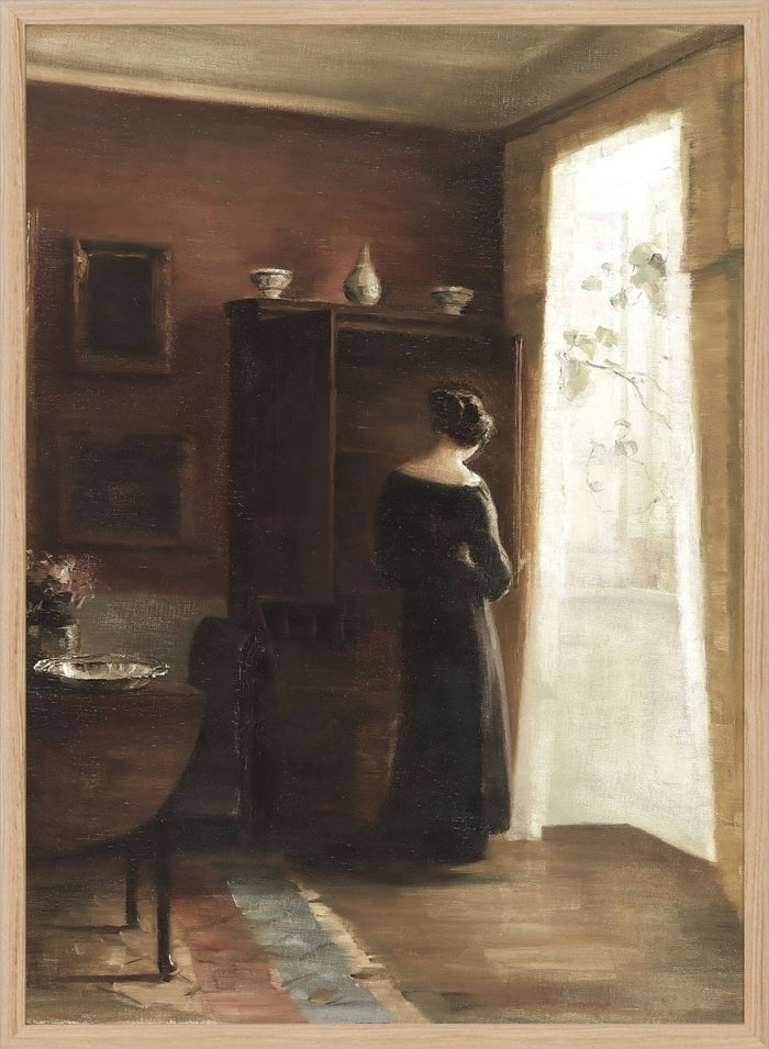 Painting of a woman standing in a sunlit room with a large mirror. Interior with a Young Girl Standing by the Window by Carl Holsøe is a vintage Danish interior art print featuring Scandinavian interiors, muted tones, and a woman by a window.