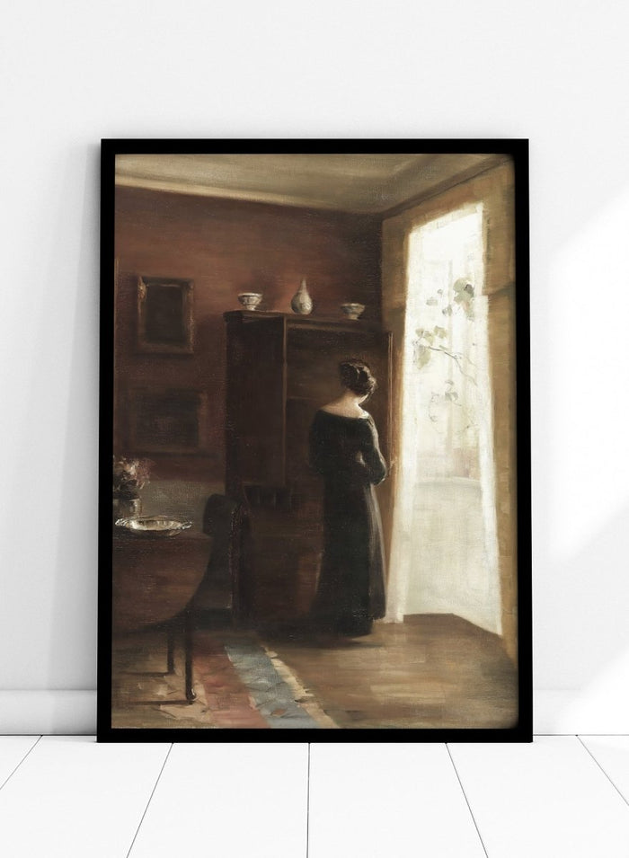 Painting of a woman standing in a sunlit room with a large mirror. Interior with a Young Girl Standing by the Window by Carl Holsøe is a vintage Danish interior art print featuring Scandinavian interiors, muted tones, and a woman by a window.