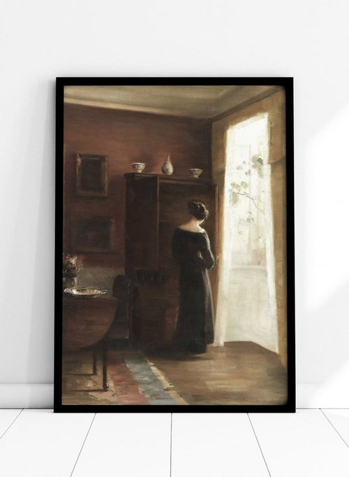 Painting of a woman standing in a sunlit room with a large mirror. Interior with a Young Girl Standing by the Window by Carl Holsøe is a vintage Danish interior art print featuring Scandinavian interiors, muted tones, and a woman by a window.