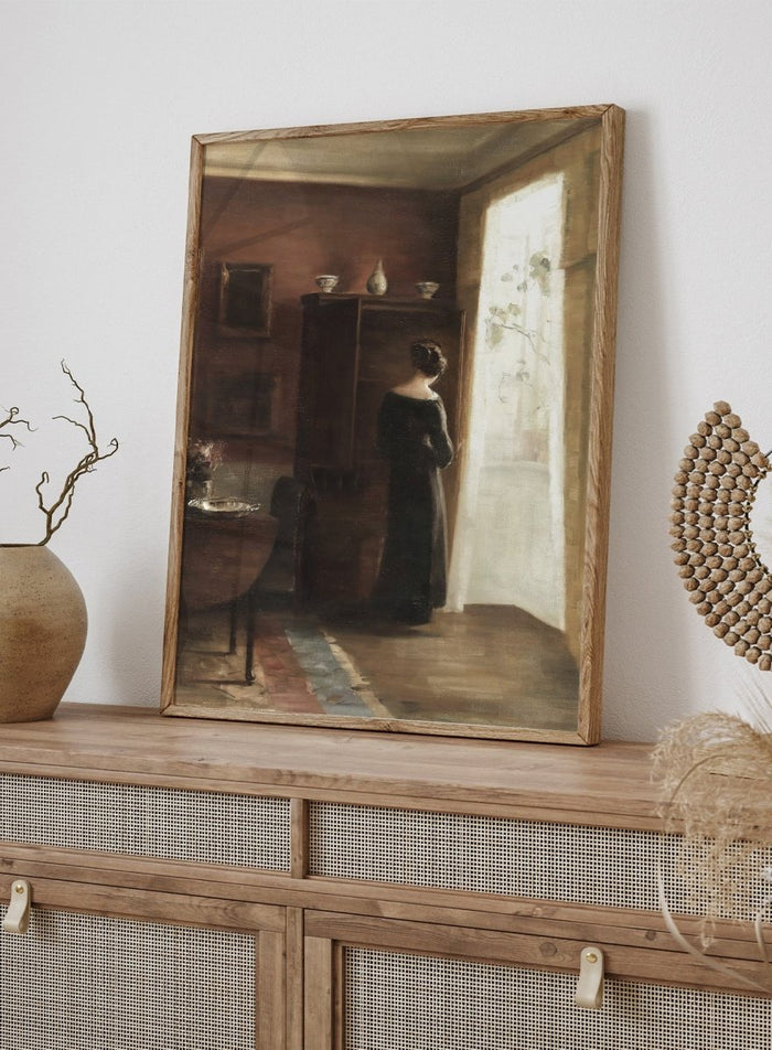 Painting of a woman standing in a sunlit room with a large mirror. Interior with a Young Girl Standing by the Window by Carl Holsøe is a vintage Danish interior art print featuring Scandinavian interiors, muted tones, and a woman by a window.
