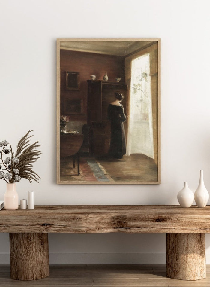 Painting of a woman standing in a sunlit room with a large mirror. Interior with a Young Girl Standing by the Window by Carl Holsøe is a vintage Danish interior art print featuring Scandinavian interiors, muted tones, and a woman by a window.