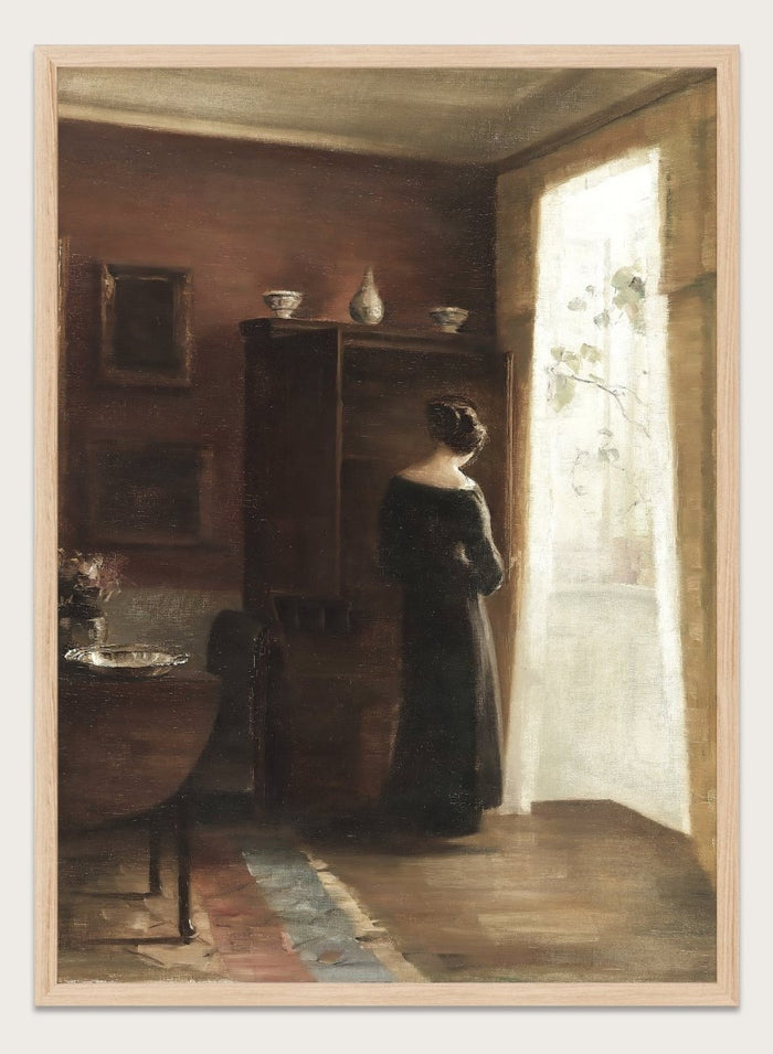 Painting of a woman standing in a sunlit room with a large mirror. Interior with a Young Girl Standing by the Window by Carl Holsøe is a vintage Danish interior art print featuring Scandinavian interiors, muted tones, and a woman by a window.