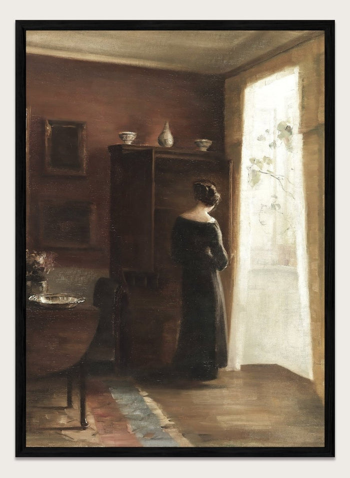 Painting of a woman standing in a sunlit room with a large mirror. Interior with a Young Girl Standing by the Window by Carl Holsøe is a vintage Danish interior art print featuring Scandinavian interiors, muted tones, and a woman by a window.