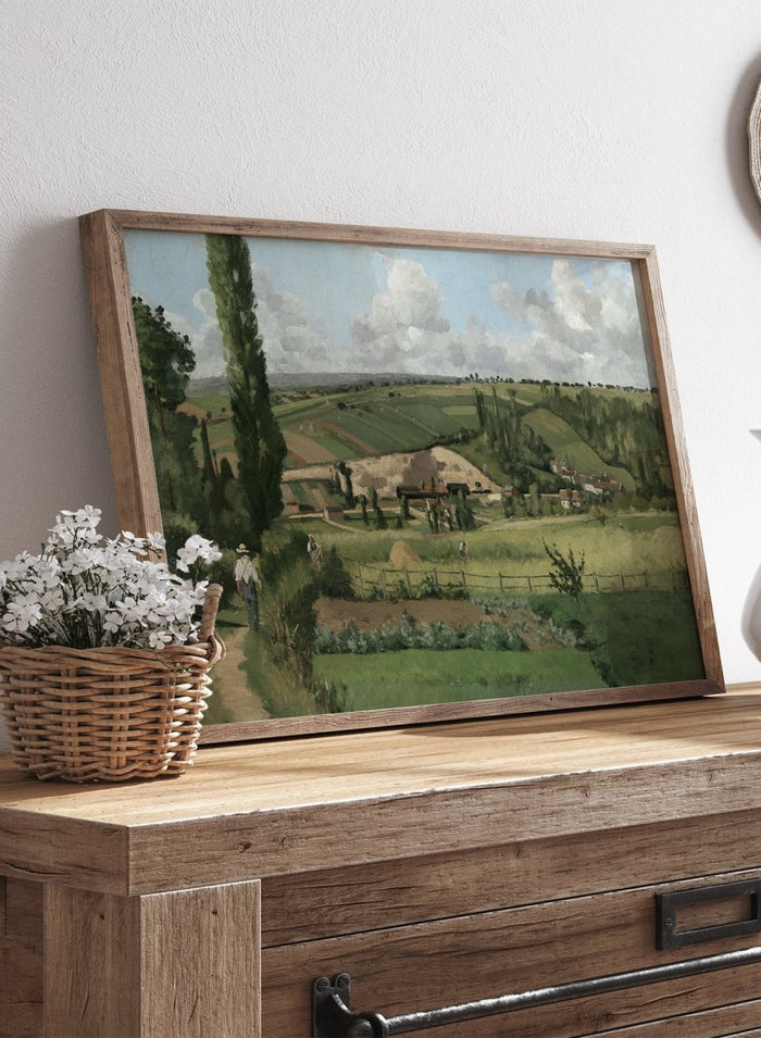A framed landscape painting depicting a green countryside scene with rolling hills, trees, a path, small buildings, and a cloudy sky. Landscape at Les Pâtis, Pontoise by Camille Pissarro is a vintage French landscape print featuring green fields, farm workers, rolling hills, and a summer sky.