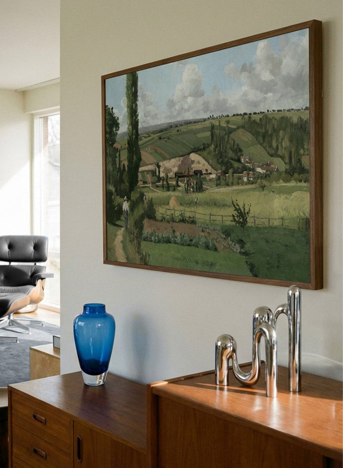 A framed landscape painting depicting a green countryside scene with rolling hills, trees, a path, small buildings, and a cloudy sky. Landscape at Les Pâtis, Pontoise by Camille Pissarro is a vintage French landscape print featuring green fields, farm workers, rolling hills, and a summer sky.