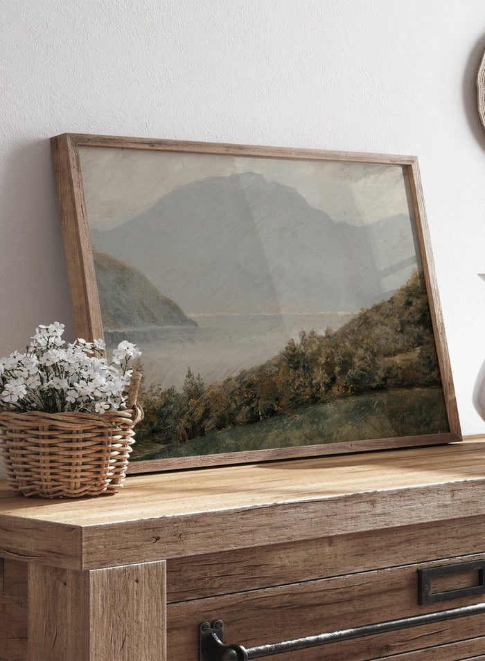 A framed landscape painting depicting a misty mountain range over a body of water with trees in the foreground. Landscape with Mountains in the Background by Susette Holten is a vintage mountain landscape print featuring a lake, misty peaks, trees, and muted natural tones.