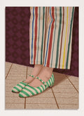 A close-up of a person's feet wearing green and white striped Mary Jane flats, paired with cream-colored striped trousers, standing on a tiled floor against a dark patterned wall. Colorful Striped Mary Janes by Lisa Solomoniuk premium framed fine art print featuring green striped shoes and colorful wide-leg trousers.