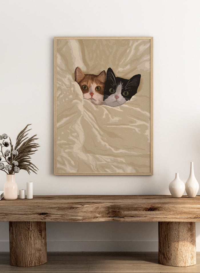 A stylized illustration of two cats peeking out from under a rumpled cream-colored blanket. The cat on the left is orange and white, and the cat on the right is black and white.  Cozy Cats Peeking Out by Lisa Solomoniuk premium framed fine art print featuring two cats tucked into soft cream bedding with wide-eyed expressions.
