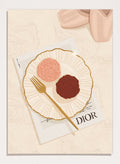 A stylized flat-lay illustration featuring a white plate with a gold rim holding two rose-shaped pastries, one light pink and one dark chocolate brown, with a gold fork on top. Elegant Cupcakes Ballet Shoes by Lisa Solomoniuk premium framed fine art print featuring pink ballet shoes, frosted cupcakes, and gold details.