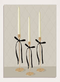 Three gold taper candle holders with cream candles and black ribbon bows, arranged on a light gray surface against a beige diamond-patterned background. Elegant Gold Taper Candles by Lisa Solomoniuk premium framed fine art print featuring gold candlesticks, cream tapers, and long black ribbons.