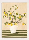 A framed still life artwork showing yellow lemons on leafy branches arranged in a white vase, set against a warm beige background with green horizontal stripes at the bottom. Lemon Tree Still Life by Lisa Solomoniuk premium framed fine art print featuring bright yellow lemons in a white vase on a striped green table.