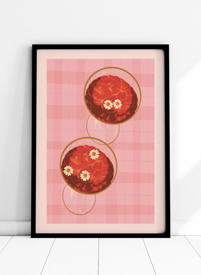 A stylized art print featuring two overlapping pink and red plaid glasses with white daisy flowers, framed by a black border on a light pink background. Pink Drinks With Daisies by Lisa Solomoniuk premium framed fine art print featuring red coupe glasses, floating daisies, and pink gingham.