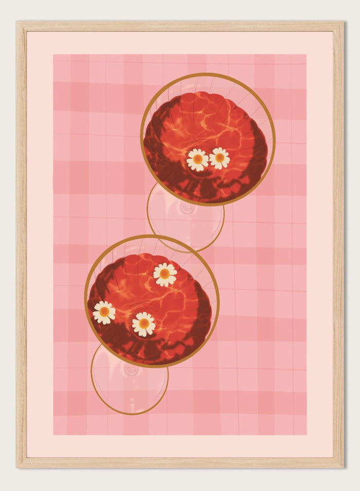 A stylized art print featuring two overlapping pink and red plaid glasses with white daisy flowers, framed by a black border on a light pink background. Pink Drinks With Daisies by Lisa Solomoniuk premium framed fine art print featuring red coupe glasses, floating daisies, and pink gingham.