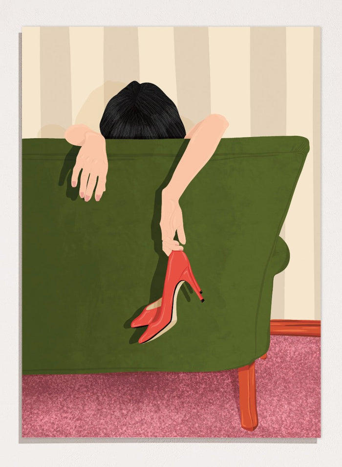 A stylized illustration of a person sitting on a green sofa, holding a pair of red high-heeled shoes. The person has dark hair and pale skin, with striped beige walls and a pink carpet in the background. Woman Resting Red Heels by Lisa Solomoniuk premium framed fine art print featuring a woman draped over a green chair holding bright red heels.