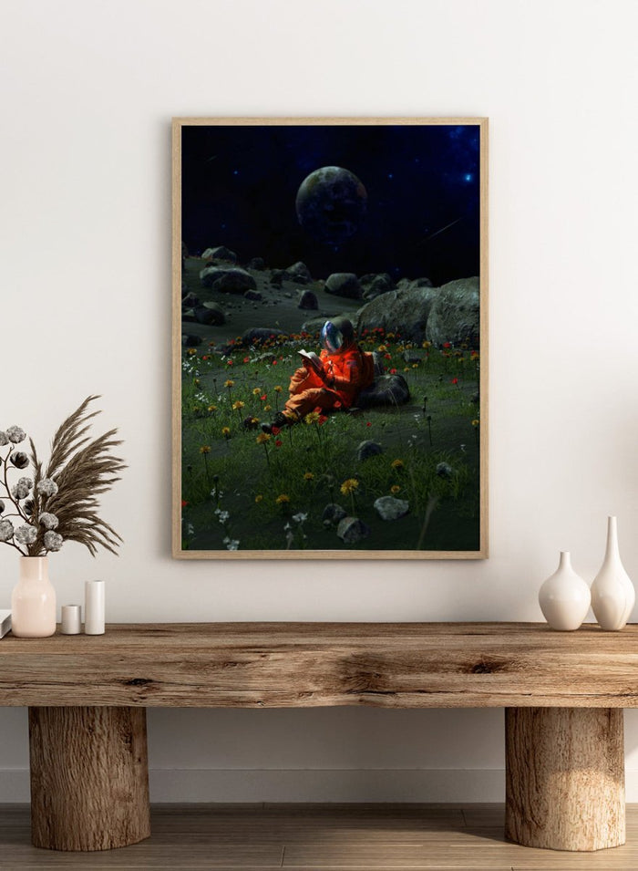 Person in orange spacesuit sitting on the moon reading a book with a large moon and stars in the background. Astronaut’s Alien Garden by Nicebleed is a surreal space art print featuring an astronaut reading in a floral alien landscape, modern aesthetic wall poster.