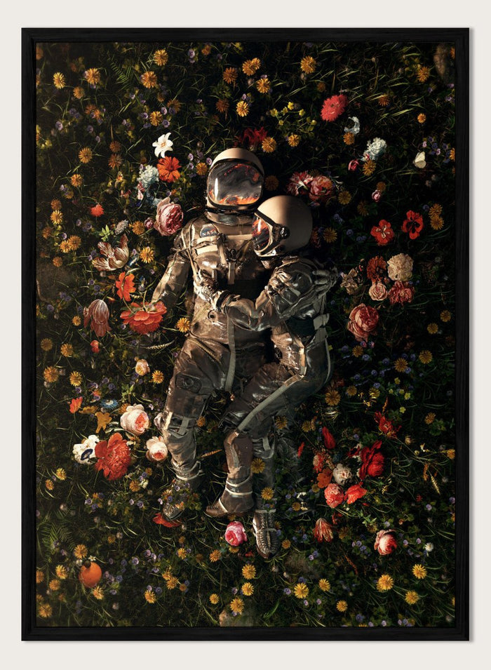 Astronaut lying on a bed of flowers with a dark background. Cosmic Couple Bloom by Nicebleed is a surreal astronaut floral art print featuring two astronauts in a field of flowers, modern aesthetic wall poster.