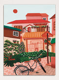 A colorful illustrated street scene with pink and peach buildings, green plants, flowers, a bicycle in the foreground, and a pale sky with a red sun. Shop Charming Village Street by Nidhi Kachhadiya, a colorful village wall art print featuring a bicycle, flower-filled homes, terracotta roofs, and warm pink and green tones.