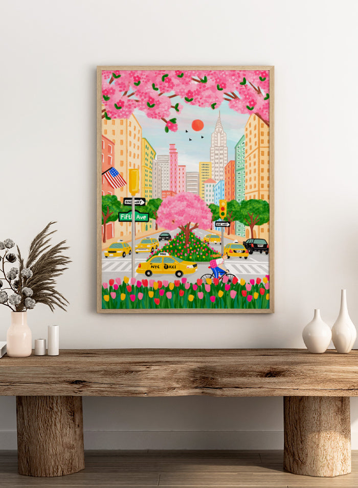 A colorful illustrated city street scene with pink cherry blossoms overhead, yellow NYC taxis, tall buildings, a bicycle with a pink basket, flowers in the foreground, and street signs including Fifti Ave. Shop NYC Cherry Blossoms by Nidhi Kachhadiya, a colorful New York City wall art print with Fifth Avenue, yellow taxis, pink blossoms, and spring city charm.
