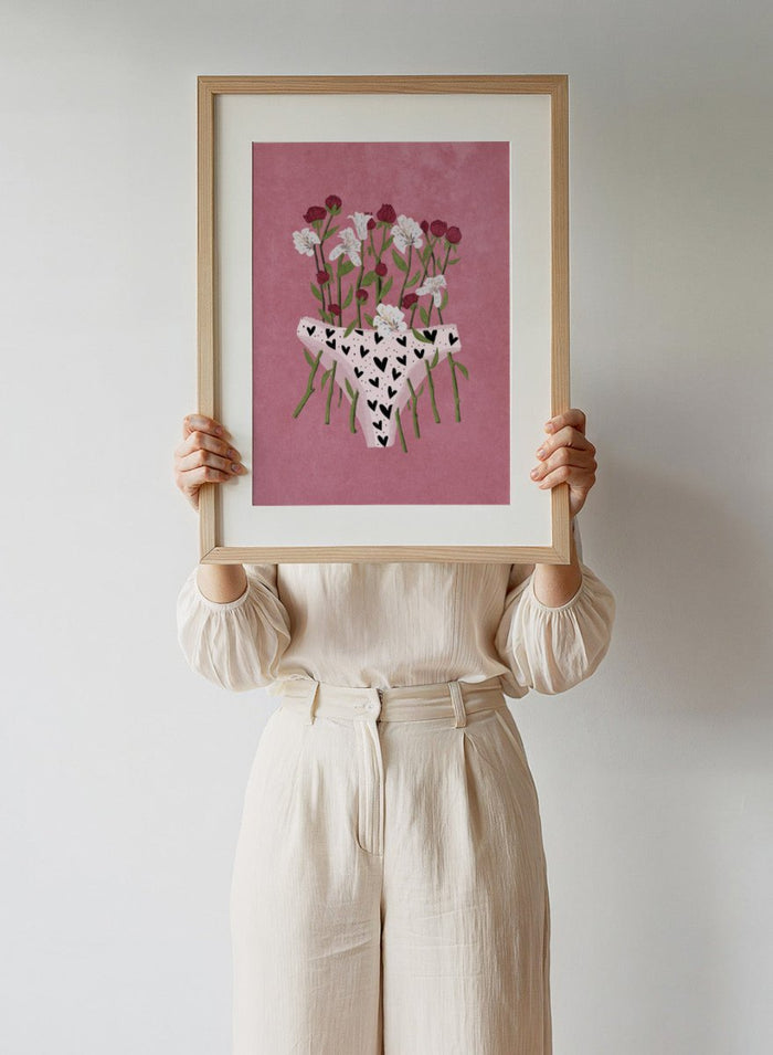 White underwear with black heart patterns surrounded by flowers on a pink background. Blooming Slip by Raissa Oltmanns features a feminist illustration of flowers growing from heart-patterned underwear on a pink background. Bold floral art print with strong feminine symbolism.