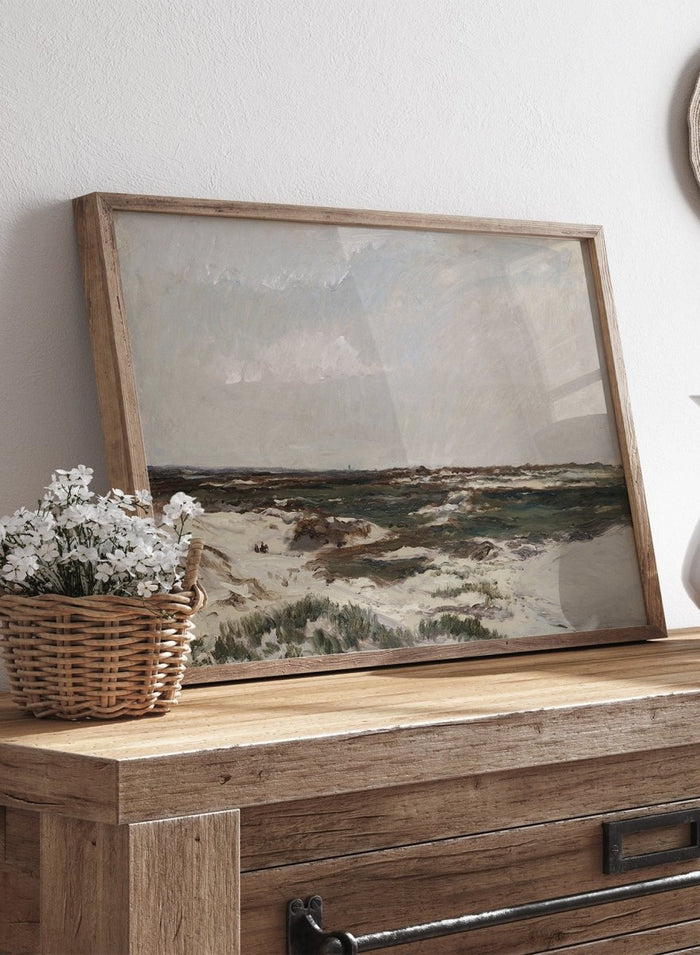 A framed landscape painting depicting a moody, overcast coastal scene with sandy dunes, dark water, and a cloudy sky. The Dunes at Camiers by Charles-François Daubigny is a vintage French coastal landscape print with sand dunes, grey skies, distant figures, and muted earth tones.