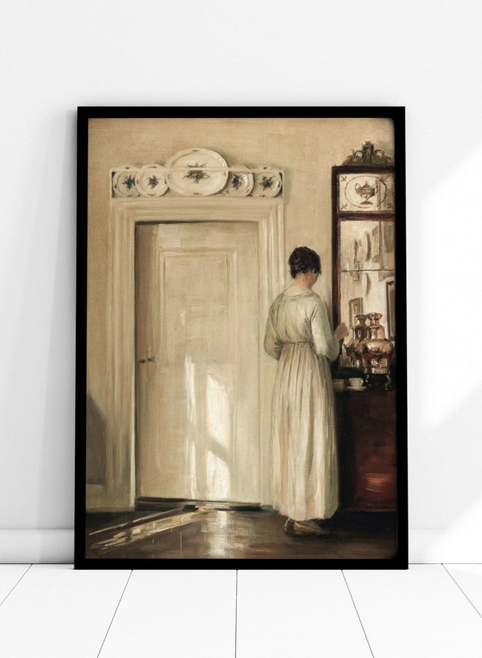 Woman in a white dress standing in a room with a door and shelves filled with items. Woman Making Tea by Carl Holsøe is a vintage Danish interior art print featuring Scandinavian home interiors, muted tones, natural light, and a woman preparing tea.