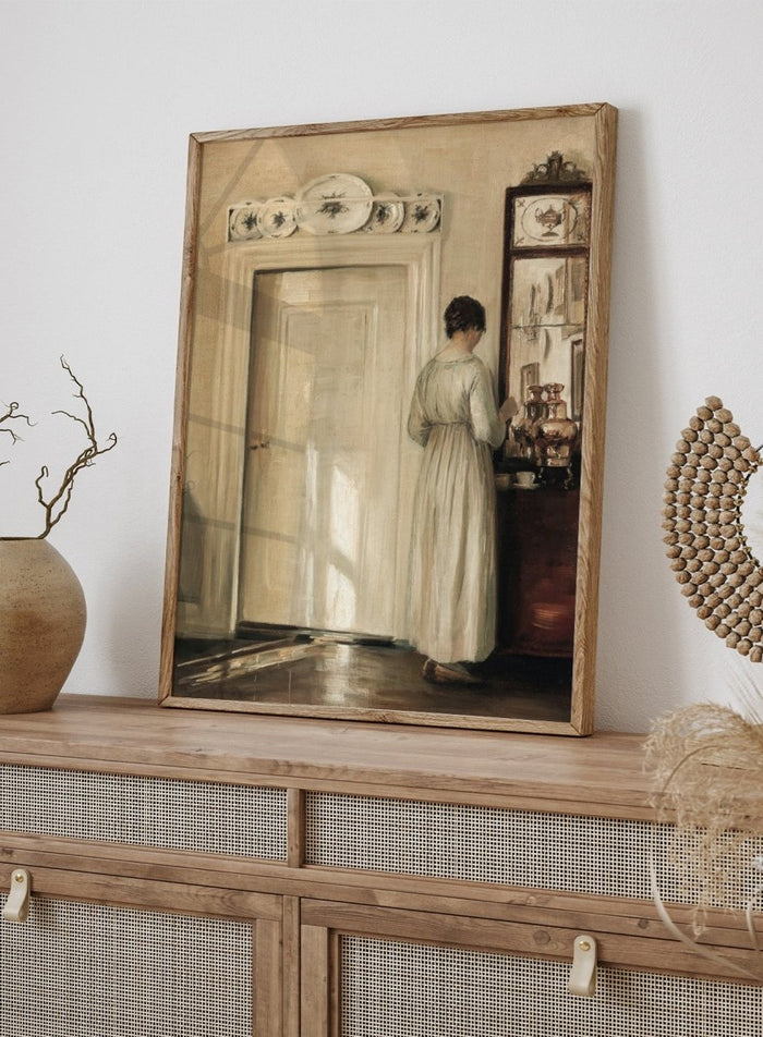 Woman in a white dress standing in a room with a door and shelves filled with items. Woman Making Tea by Carl Holsøe is a vintage Danish interior art print featuring Scandinavian home interiors, muted tones, natural light, and a woman preparing tea.