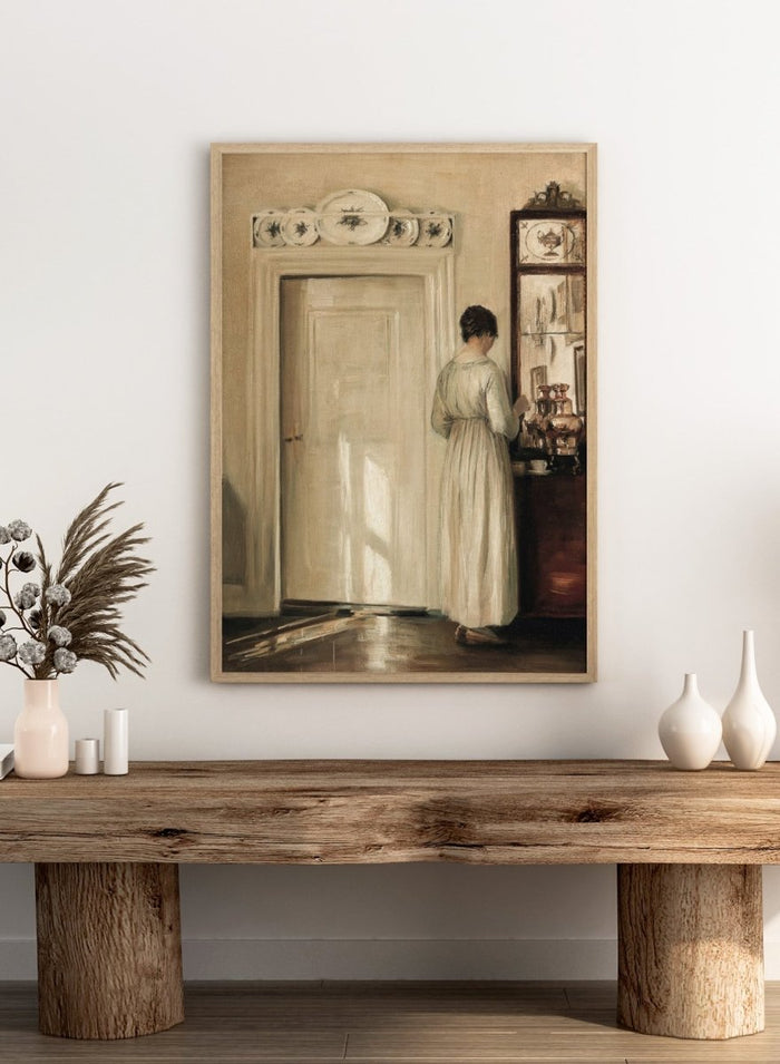 Woman in a white dress standing in a room with a door and shelves filled with items. Woman Making Tea by Carl Holsøe is a vintage Danish interior art print featuring Scandinavian home interiors, muted tones, natural light, and a woman preparing tea.