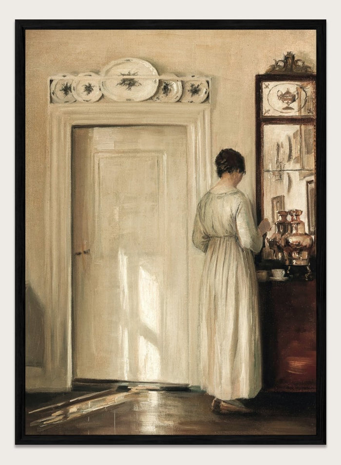 Woman in a white dress standing in a room with a door and shelves filled with items. Woman Making Tea by Carl Holsøe is a vintage Danish interior art print featuring Scandinavian home interiors, muted tones, natural light, and a woman preparing tea.